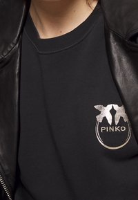 Felpa nera con scollatura rotonda, caratterizzata da un logo argento e un dettaglio di design sul lato sinistro del petto. Realizzata in tessuto morbido; indossata con una giacca di pelle.