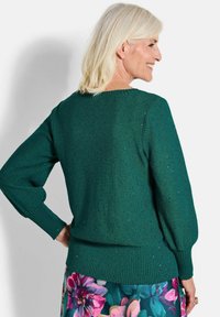 Grüner gestrickter Pullover mit geripptem Saum und Bündchen, der ein strukturiertes Muster und leichten Glitzer zeigt. Über einem Rock mit floralem Print getragen.