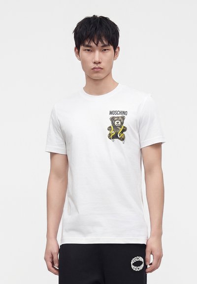 Νέος άνδρας φοράει λευκό t-shirt Moschino με γραφικό αρκουδάκι και μαύρο σορτς με το λογότυπο Moschino στο αριστερό πόδι.