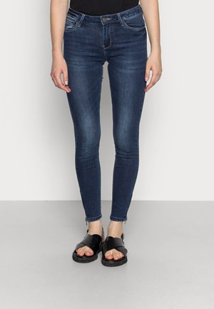 Femme portant un jean skinny bleu foncé et des sandales noires à bouts ouverts, debout devant un fond clair uni.