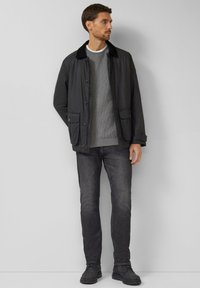 Uomo con giacca nera sopra maglione grigio, jeans scuri e scarpe nere, in piedi davanti a uno sfondo grigio chiaro uniforme.