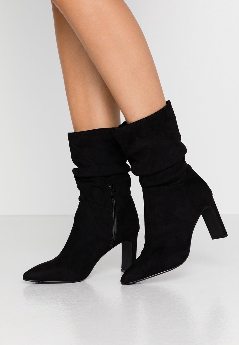 Femme portant des bottes mi-mollet en daim noir décontractées avec fermeture éclair latérale et talons blocs, debout sur un sol blanc lisse.