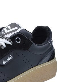 Deportivas negras con diseño perforado, cordones blancos y suela de goma. Presenta un parche con el logo y acentos texturizados en la lengüeta y el talón.