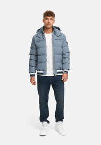 Chaqueta acolchada azul claro con capucha, que cuenta con bolsillos con cremallera, puños acanalados y un dobladillo a rayas; usada sobre una camiseta blanca y jeans oscuros.