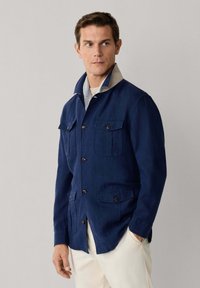 Mand iført marineblå jakke med knapper og fire lommer med klap samt beige krave, kombineret med lyse bukser, stående foran en ensfarvet baggrund.