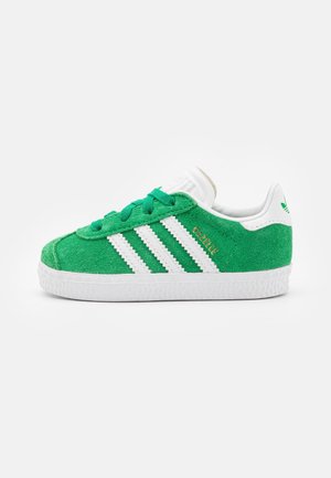 Groene suède sneaker met een witte rubberen zool en drie witte strepen. Voorzien van groene veters en een witte gewatteerde kraag. Gouden tekst leest "Gazelle."