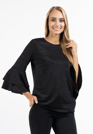Femme aux longs cheveux blonds souriante, portant un haut noir pailleté avec des manches cloche en couches et un pantalon noir, sur fond blanc.