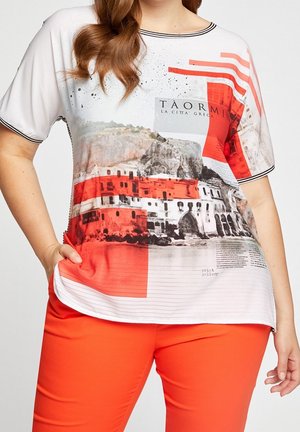 Frau trägt ein kurzärmeliges Top mit einem rot-weißen Motiv von Gebäuden in Taormina und leuchtend orangefarbene Hosen, die Hand in der Tasche.