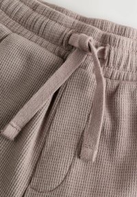 Svetlohnedé rebrované tepláky s pásom na stiahnutie, prednými vreckami a textúrovanou tkaninou. Obsahuje jemný, tkaný vzor.