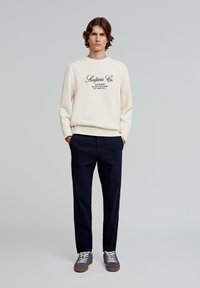 Lichtcrème sweatshirt met zwart tekstlogo, donker marineblauwe broek en grijze sneakers met witte veters. Eenvoudige, casual outfit met een relaxte pasvorm.