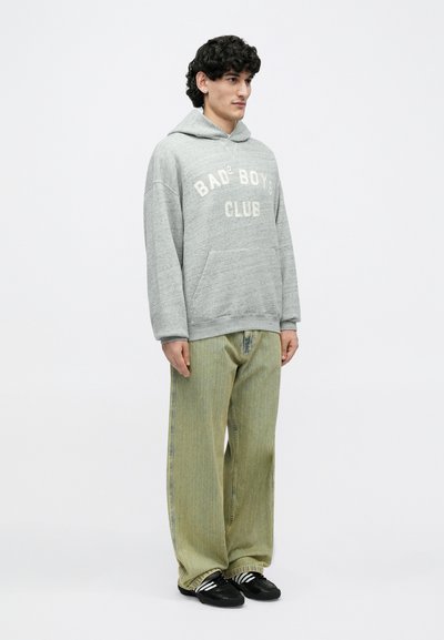 Uomo con capelli ricci e scuri che indossa una felpa grigia con la scritta "Bad Boys Club", jeans verdi larghi, calzini a righe e scarpe da ginnastica nere, in piedi su sfondo bianco.