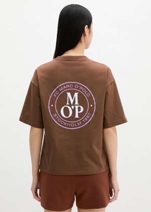 Άτομο με μακριά μαύρα μαλλιά, φορώντας καφέ μπλουζάκι Marc O’Polo με μωβ λογότυπο και κείμενο, συνδυασμένο με καφέ σορτς, με την πλάτη γυρισμένη.