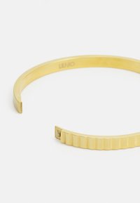 Gullfarget metallmanchettarmbånd med en glatt overflate og en ribbet seksjon i den ene enden. Inngravert logo på innsiden av båndet.