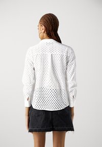 Blusa bianca con motivo ad occhio di pernice, maniche lunghe e colletto abbottonato, abbinata a una gonna in denim nera. La vista posteriore mostra i dettagli del tessuto.