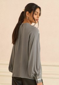 Šedá blůza s volným střihem, zvonovými rukávy a knoflíkovými detaily na manžetách. Nabízí klíčovou dírku na zadní straně a hladký materiál.
