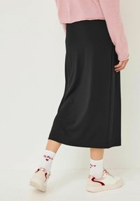 Personne portant une jupe midi noire, des baskets blanches avec des accents en or rose, des chaussettes blanches avec un texte rouge, et un pull rose, se tenant sur un sol blanc.