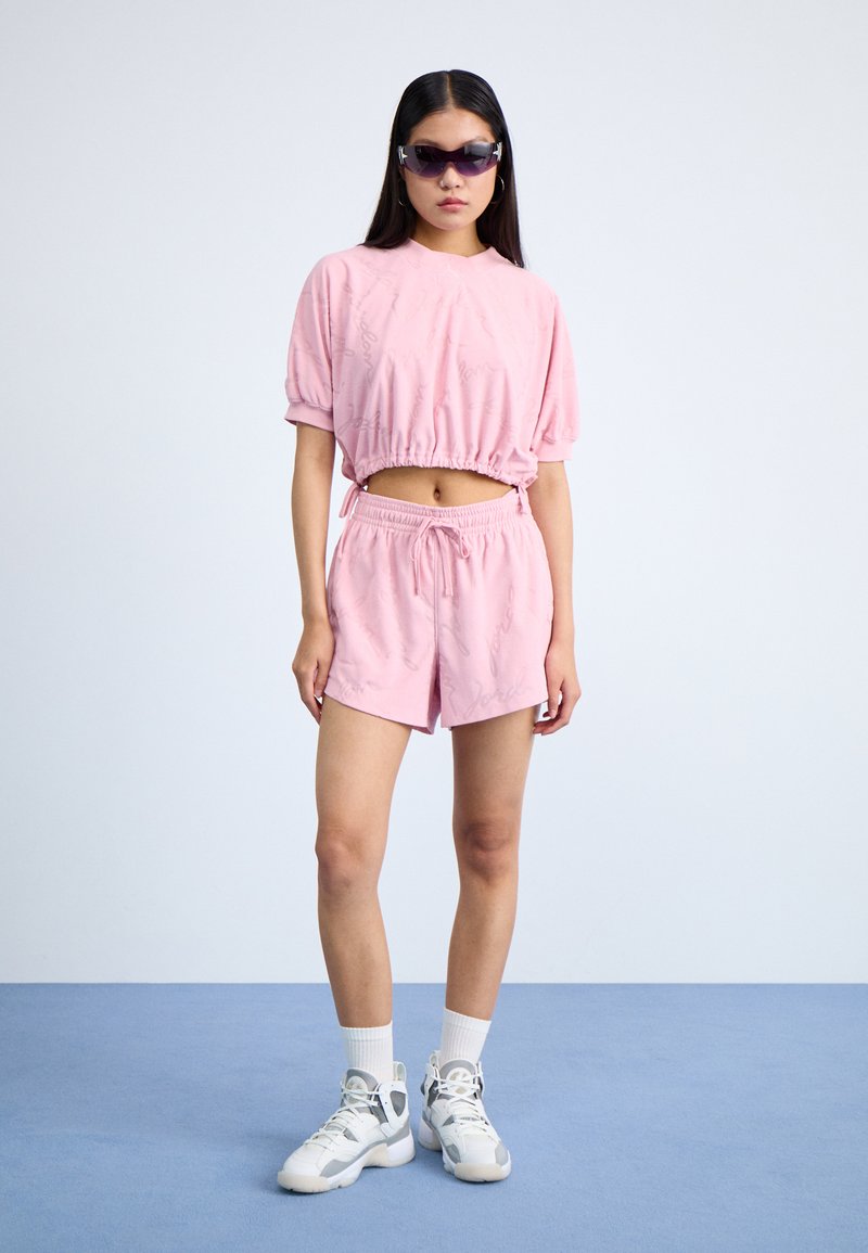 Top corto rosa con vita arricciata e pantaloni corte abbinati. Entrambi presentano delicati motivi testuali. Il modello indossa sneakers bianche e occhiali da sole scuri.