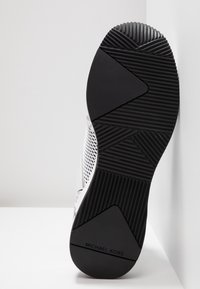 Semelle d'une chaussure de sport noire et blanche avec grip texturé et logo Michael Kors au talon, positionnée verticalement contre une surface blanche.
