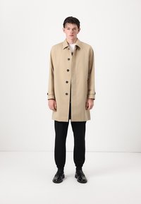 Beige trench coat with black buttons, long sleeves, and slight flare at the bottom, worn over black pants and shoes, set against a white background.