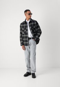 Svarte og hvite rutete skjorte, knappet med to frontlommer, båret over en hvit skjorte. Grå jeans og svarte sko fullfører antrekket.