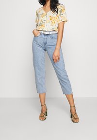 Blouse à manches courtes florale de couleur jaune clair avec des accents orange et verts, associée à un jean court bleu clair et des talons verts à lanières.