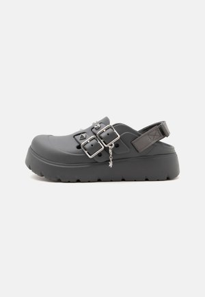 Grå slip-on træsko med to sølvspænder, perforeret overdel og en tyk, ribbet sål.