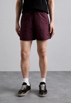 Bordeaux shorts med teksturert overflate, elastisk midjebånd og snor. Kombinert med svarte joggesko og hvite ankelsokker.