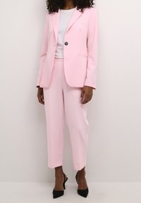 Lichtroest op maat gemaakte pak bestaande uit een blazer en cropped broek, gecombineerd met een witte blouse. Zwarte hoge hakken maken de look compleet.