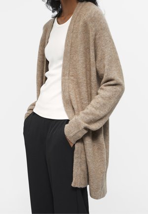 Frau trägt eine beige, offene Strickjacke über einem weißen Hemd und schwarzen Hosen, mit den Händen in den Taschen, vor einem neutralen Hintergrund.