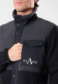 Giacca in fleece nera e grigia, con colletto alto, bottoni a pressione e taschino sul petto con logo. Texture morbida, vestibilità rilassata.