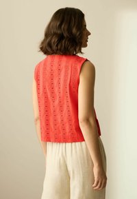 Haut rouge sans manches avec motif ajouré, design court, et surface texturée, associé à un pantalon beige clair. Style simple et décontracté.