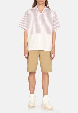 Camicia a maniche corte con strisce verticali bordeaux su fondo bianco; abbinata a pantaloni corti beige e sneakers alte bianche.