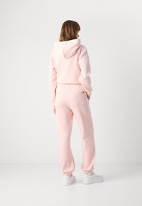 Γυναίκα γυρισμένη με φως ροζ φούτερ και ταιριαστές φόρμες με λευκά αθλητικά παπούτσια, μπροστά σε απλό λευκό φόντο.