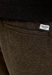 Bruna tygbyxor med texturerad yta, med en liten vit logotagg märkt "JACK & JONES" nära fickan.