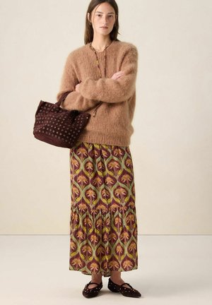 Woman stands with arms crossed wearing a fuzzy brown sweater, patterned long skirt, dark studded handbag, and matching studded flats.