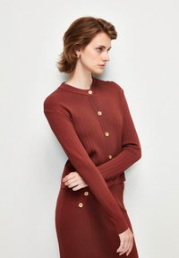 Cardigan lavorato a maglia color ruggine con colletto tondo, texture a coste e bottoni dorati sul davanti. Design attillato con maniche lunghe.