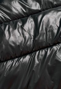 Melnā polsterētā auduma ar spīdīgu tekstūru, kurā ir šūtas sadaļas un lielas, daļēji caurspīdīgas burti, kas iemontēti materiālā.