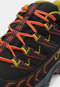 Černá sportovní bota s vícebarevnými žlutými a červenými tkaničkami. Texturovaný svrchní materiál; viditelné výrazné logo a vzorovaná podrážka.