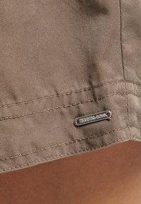 Bruine stof met een gestructureerd oppervlak en zichtbare stiknaden. Metalen logoplek is bevestigd nabij de zoom.