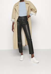 Beige Trenchcoat, hellblau gestreiftes Rippenoberteil, schwarze Lederhose und weiße Sneaker. Der Mantel hat einen Gürtel und weite Ärmel.