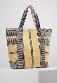 Borsa a tracolla intrecciata con motivi a strisce gialle, nere e bianche, dotata di due manici robusti e tessuto testurizzato. Design leggermente strutturato.