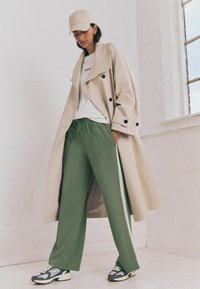 Beige trench coat s černými knoflíky, bílý tričko s nápisem "PARIS", zelené široké tepláky a šedé tenisky.