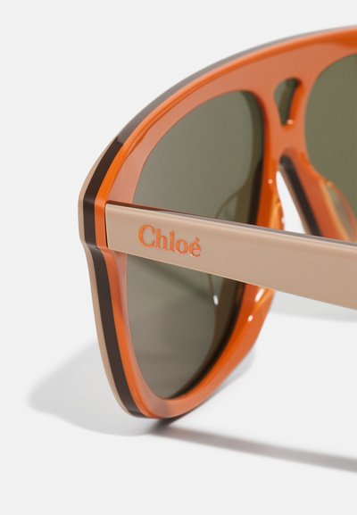 Lunettes Chloé avec une monture audacieuse orange et beige, dotées de verres vert foncé et d'une texture lisse, mises en valeur par un détail de marque.