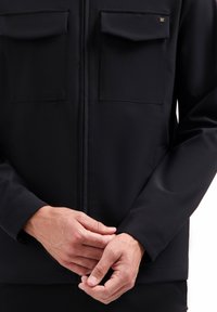 Man in een zwarte jas met klepzakken, handen voor zich gevouwen, tegen een witte achtergrond.