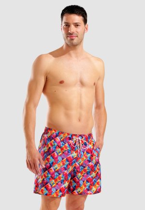 Hemdloser Mann steht mit Händen in der Nähe von bunten Badeshorts mit geometrischem Muster vor einem schlichten hellen Hintergrund.