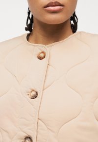 Gros plan sur une personne portant une veste matelassée beige avec de grands boutons en écaille de tortue et un col rond.