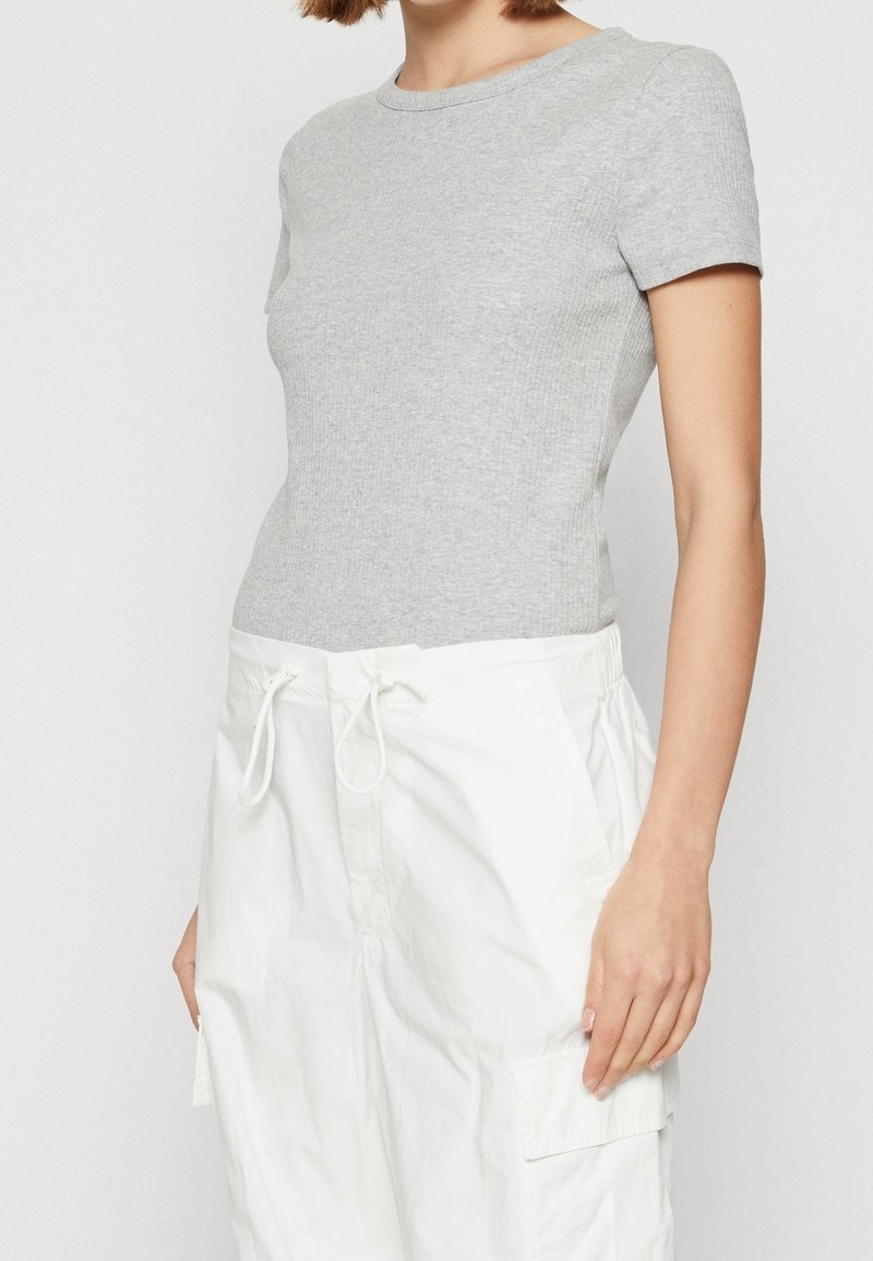 Personne portant un t-shirt gris côtelé à manches courtes ajusté, associé à un pantalon blanc ample avec cordon de serrage et poches latérales, debout devant un fond uni.