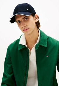 Gorra de marinero con detalles en blanco, chaqueta verde y polo blanco. La chaqueta tiene un cierre con cremallera y un pequeño logo en el pecho.