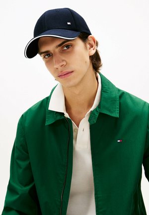 Casquette marine avec des accents blancs, veste verte et polo blanc. La veste a une fermeture zippée et un petit logo sur la poitrine.