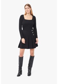 Schwarzes Langarmoberteil mit quadratischem Ausschnitt, kombiniert mit einem schwarz gefältelten Rock, der an der Seite drei silberne Knöpfe hat. Schwarze kniehohe Stiefel.